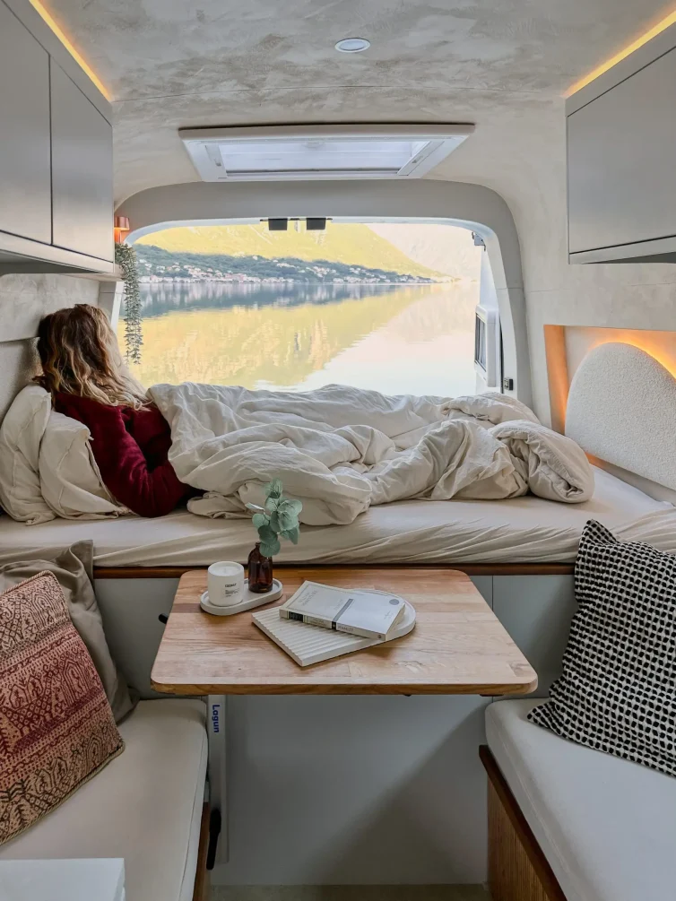 Interior view of a campervan featuring a bed, table, and scenic window view of mountains and water.